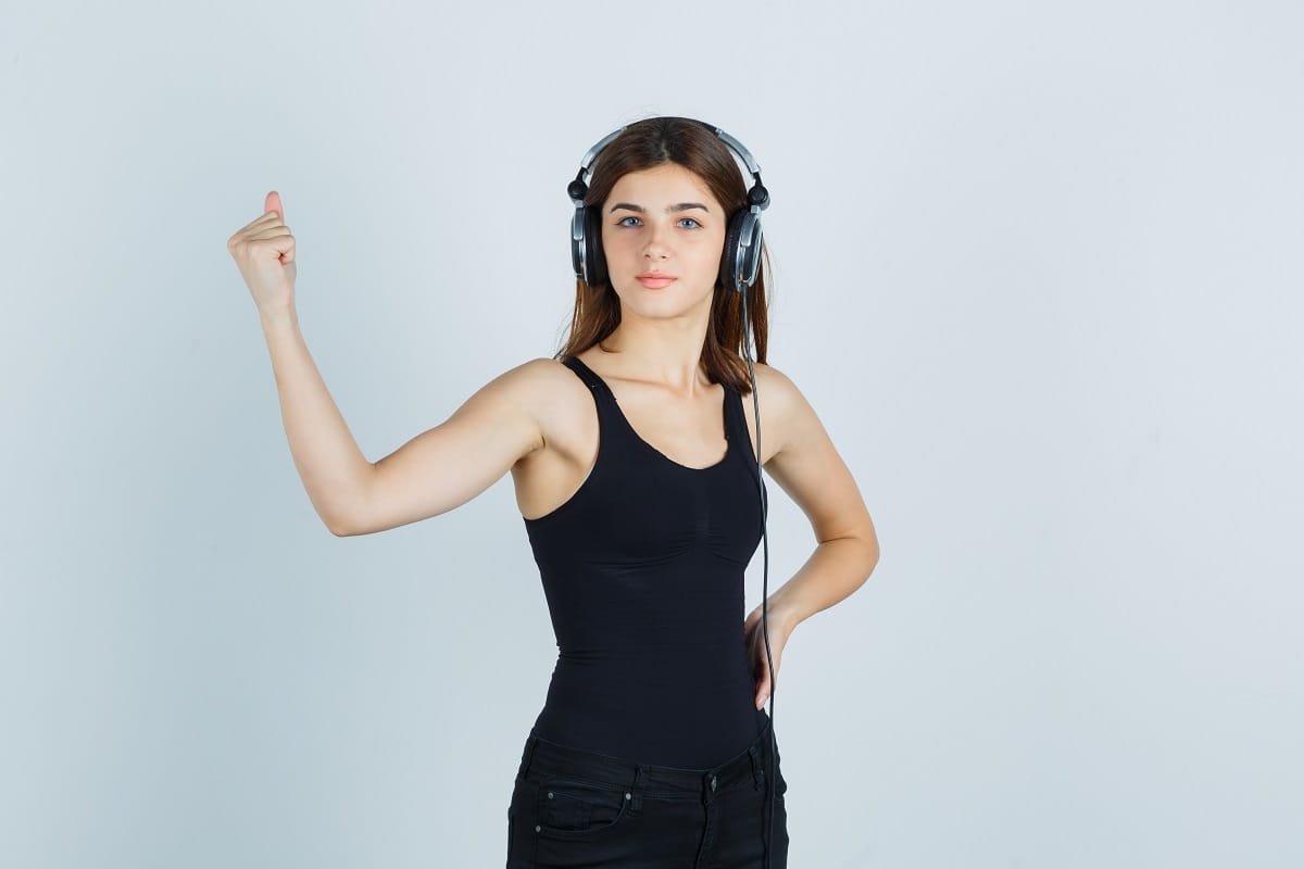 young lady listening to music with headphones while showing muscles of arm in tank top, pants and looking confident. front view. welcome to audiopick.in
